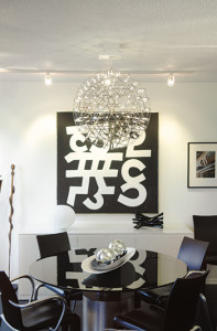 In the dining room, black chairs by French designer Philippe Starck surround a Vignelli table, which features a gray glass  top and silver leaf base. A Moooi Raimond  light fixture filled with LED lights hangs over the table. The art on the wall is an acrylic on canvas by internationally recognized artist Ulfert Wilki.